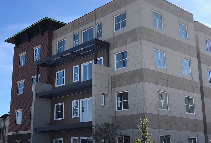 Modern senior living building exterior with brick accents, light siding, balconies, and many windows.