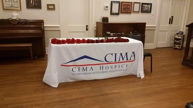 Table with CIMA Hospice banner and red roses indoors, in a wood-floored room.