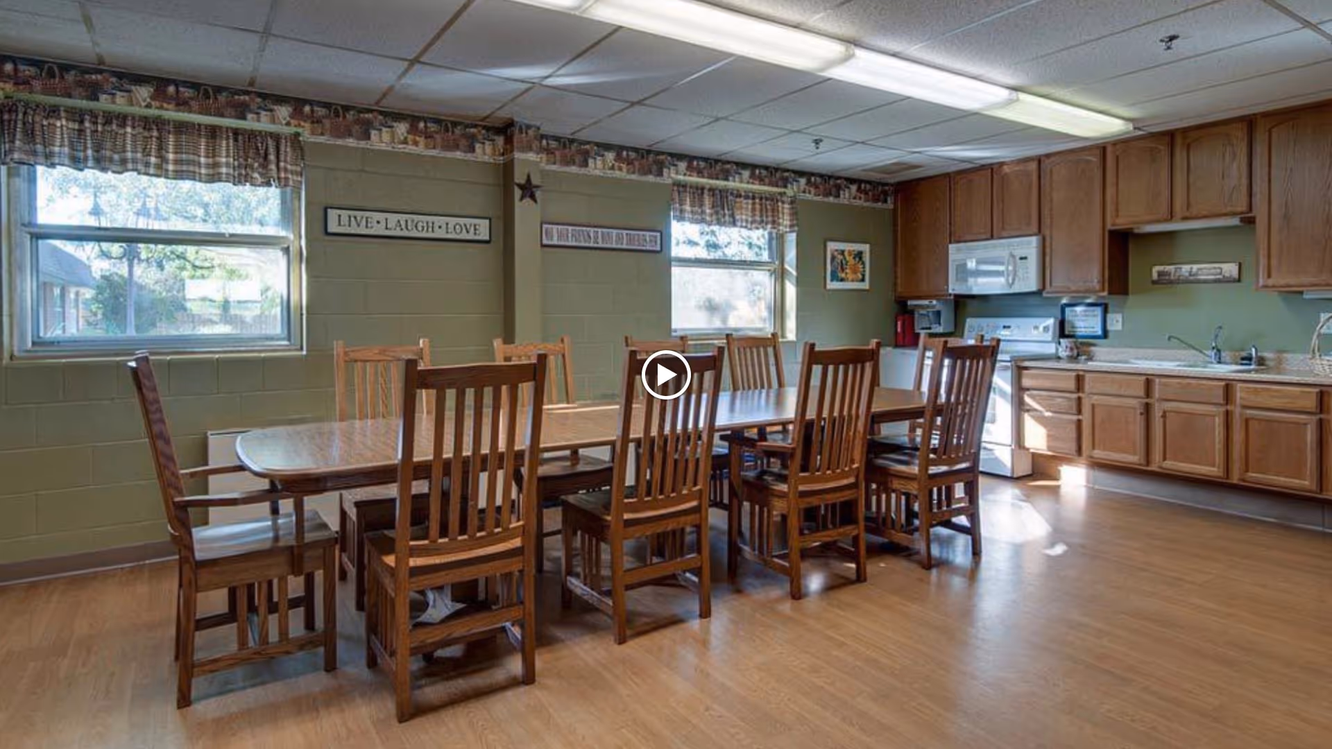A dining area in Hilltop Manor featuring a long wooden table surrounded by eight wooden chairs. The room has two windows with plaid valances, green walls with decorative signs including one that says 'LIVE • LAUGH • LOVE', and a kitchen area with wooden cabinets, a white stove, microwave, and sink. The floor is light wood, and the ceiling has fluorescent lighting.