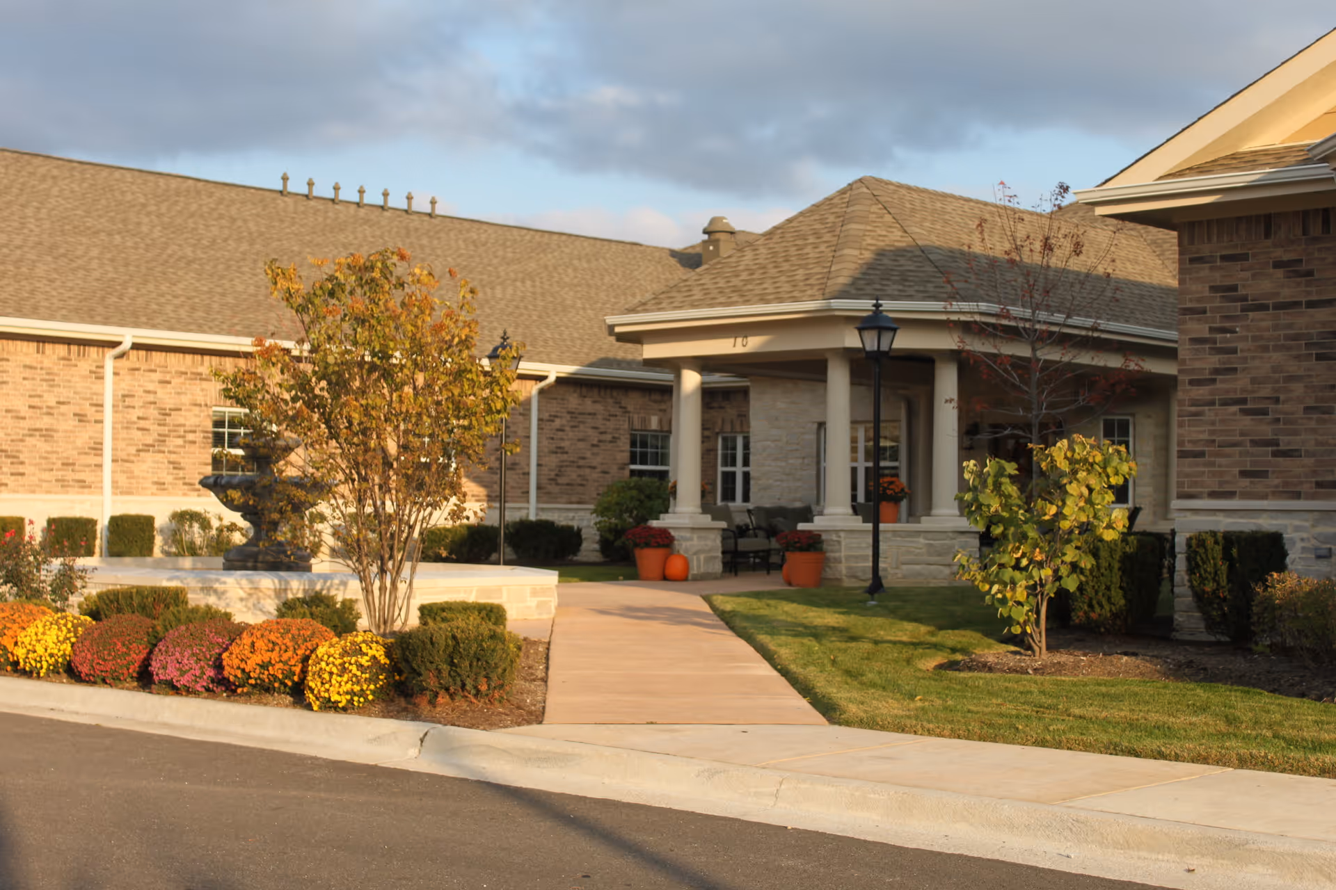 Exterior view of a senior living facility with a brick facade, a covered entrance supported by columns, a paved walkway, landscaped bushes, colorful flowers, and small trees under a partly cloudy sky.
