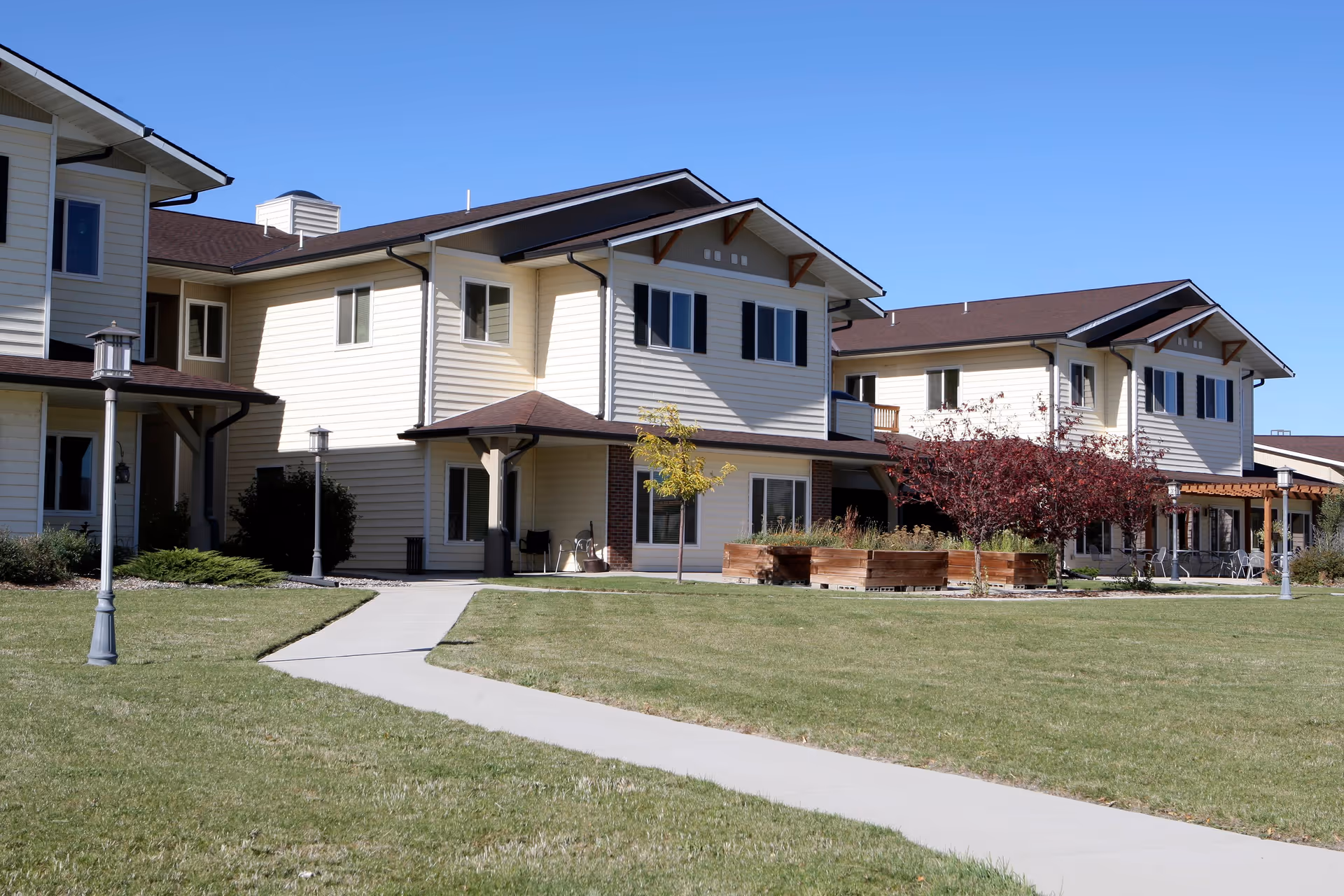 Exterior view of a two-story senior living facility building with beige siding and brown roof. There is a paved walkway leading through a well-maintained grassy lawn with lamp posts, small trees, and raised garden beds. The sky is clear and blue.