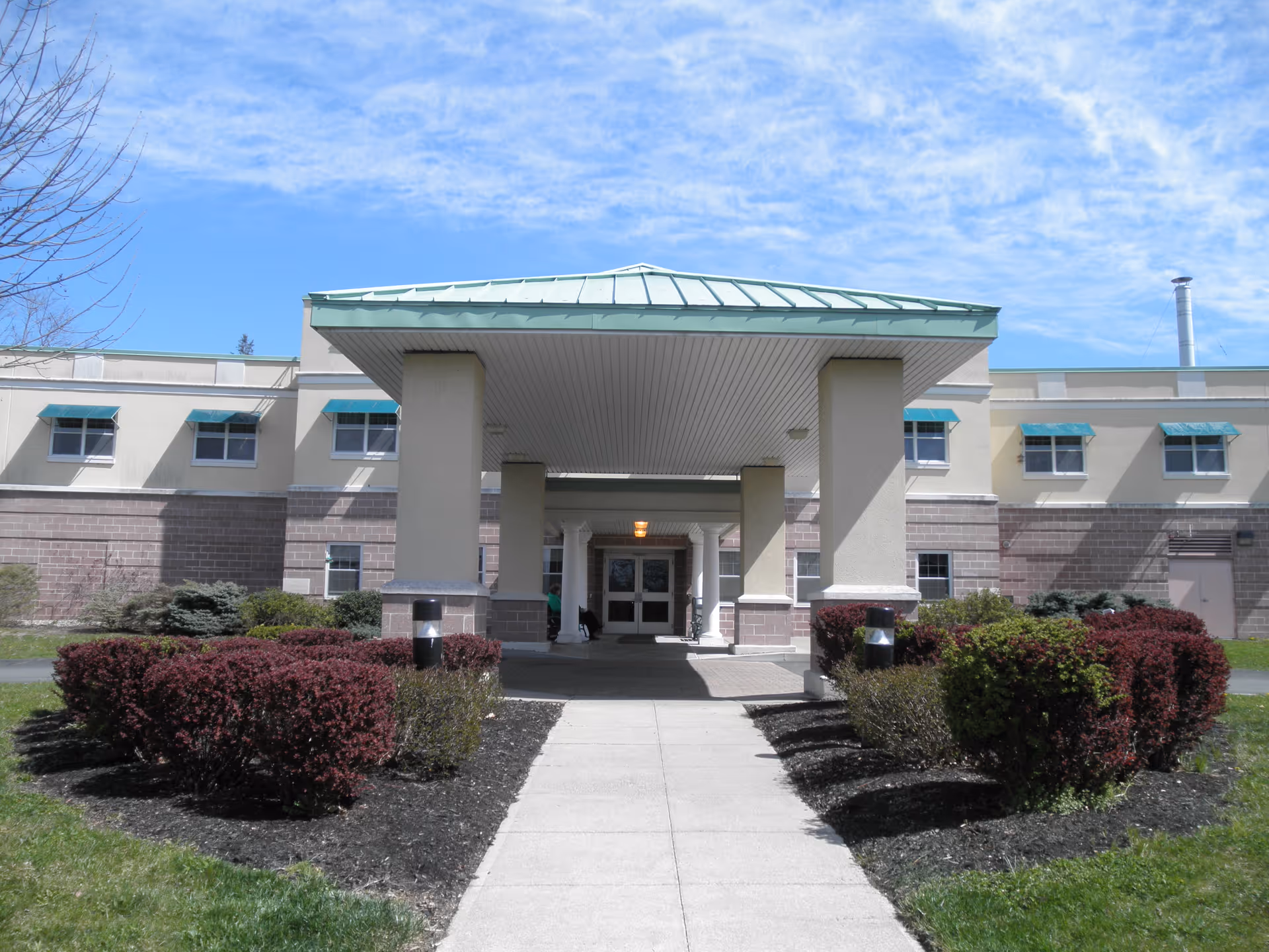 Front entrance of a rehabilitation and nursing facility with a covered drop-off area supported by large columns, surrounded by neatly trimmed bushes and a clear blue sky overhead.