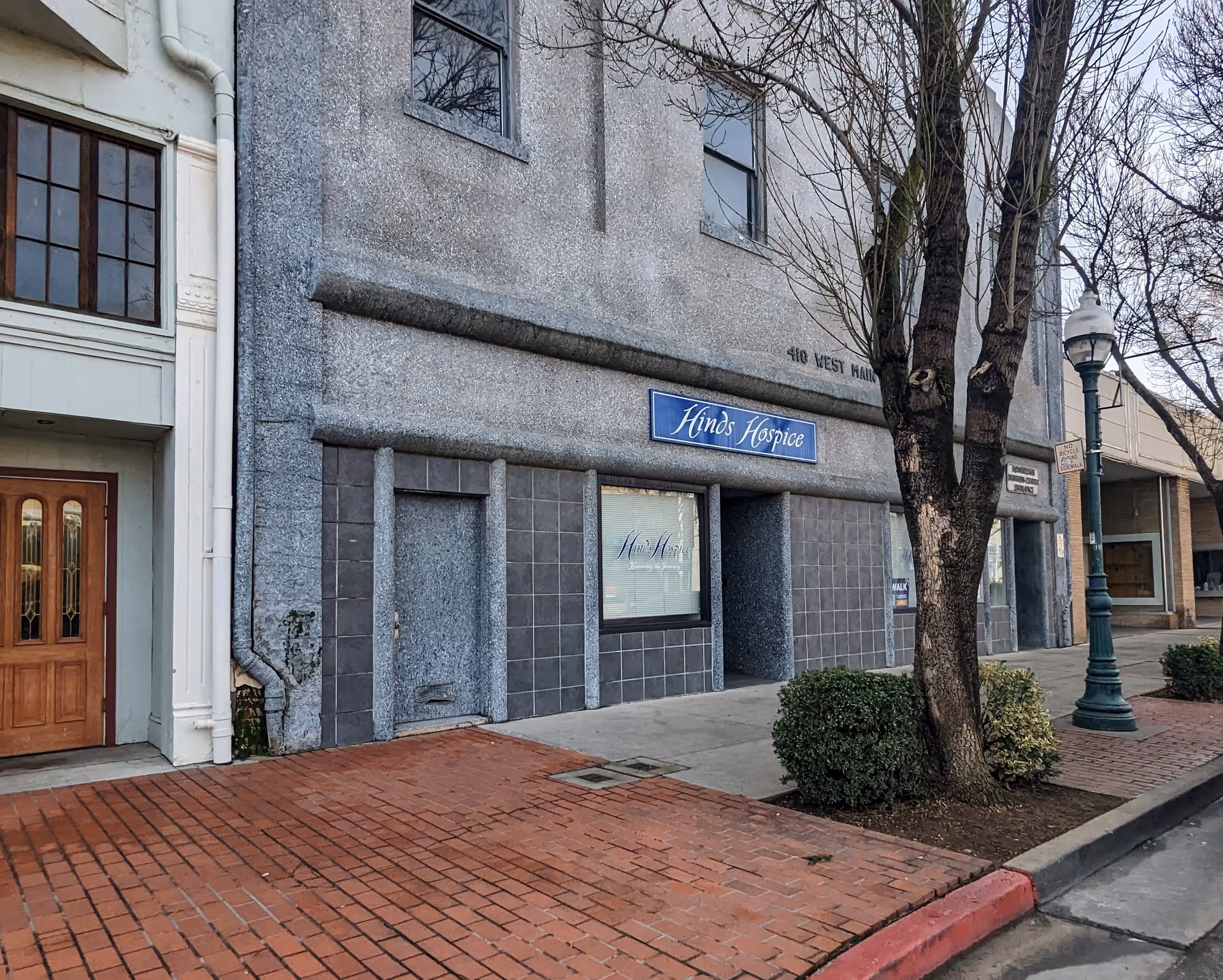 Exterior view of a grey building with a blue Hinds Hospice sign along brick sidewalk.