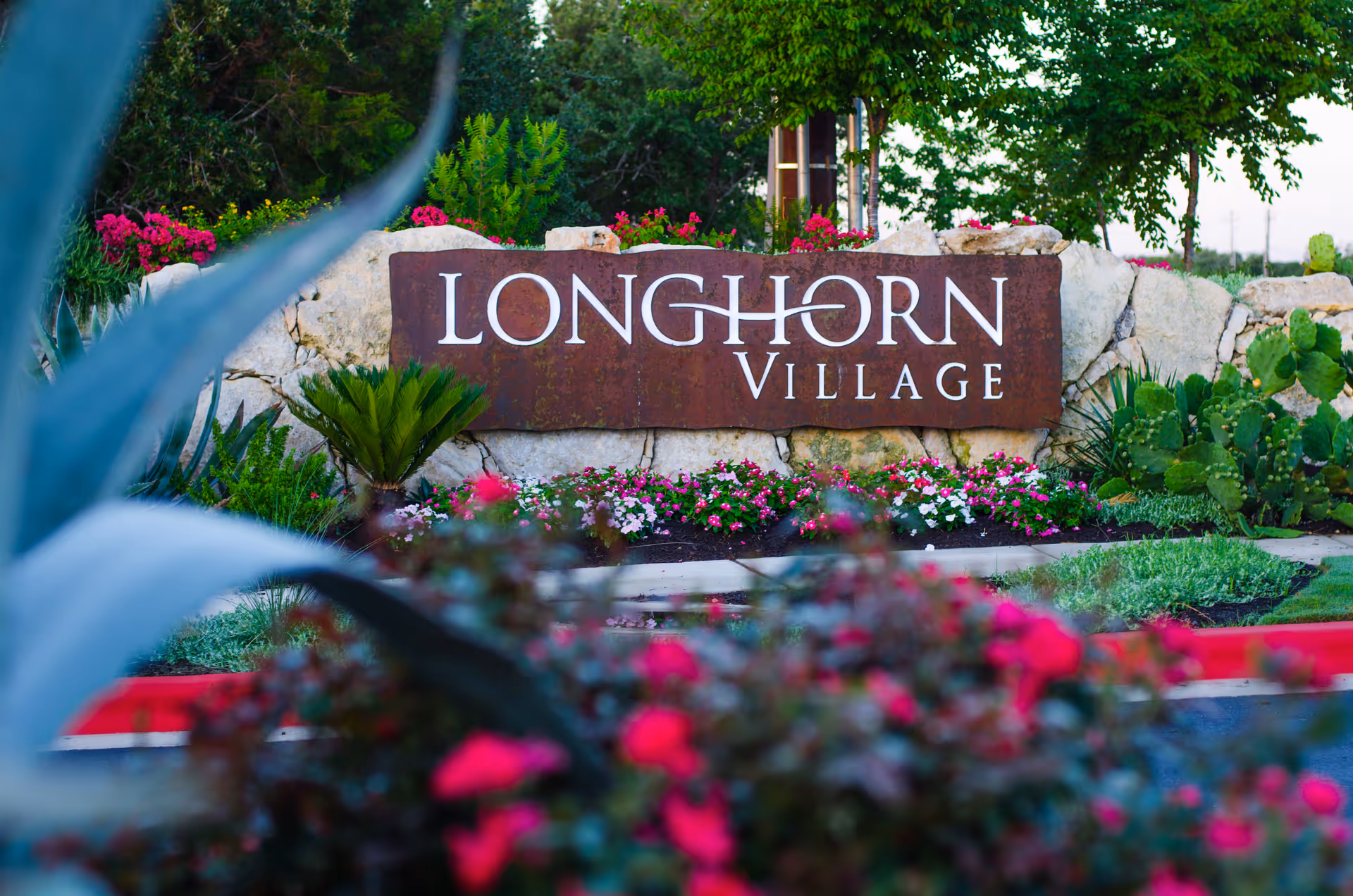 Rust-colored Longhorn Village sign framed by rocks and flowering landscaping at entrance.