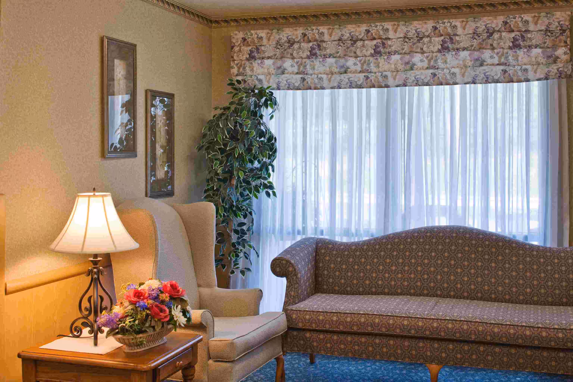 Cozy living room with a patterned sofa, armchair, lamp, flowers, and a large window.