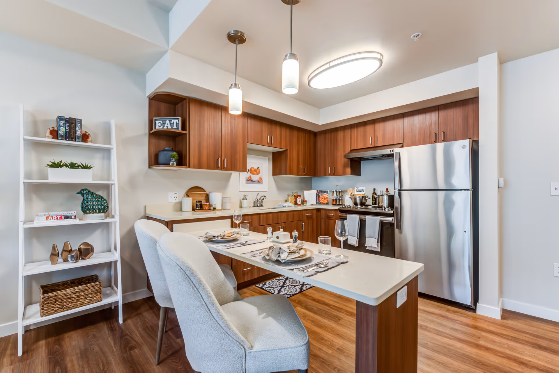 Modern kitchen with wooden cabinets, stainless steel refrigerator and stove, a white countertop island set for two with plates, glasses, and cutlery, and a white shelving unit with decorative items on the left.