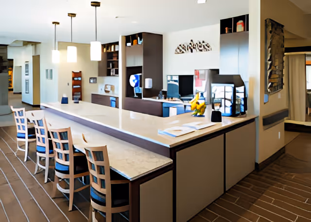 Interior view of a modern reception or common area with a long marble countertop and several wooden chairs with blue cushions lined up along it. The background includes built-in shelves, a computer monitor, decorative items, and pendant lights hanging from the ceiling. The floor has a striped carpet pattern and the walls are painted in neutral tones.
