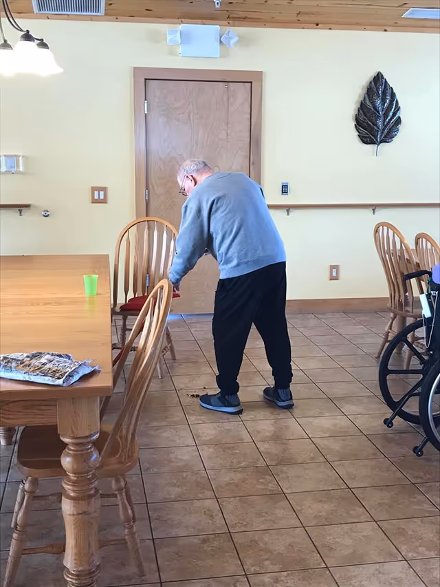 Elderly man bends over near a wooden dining table in a bright senior living dining room