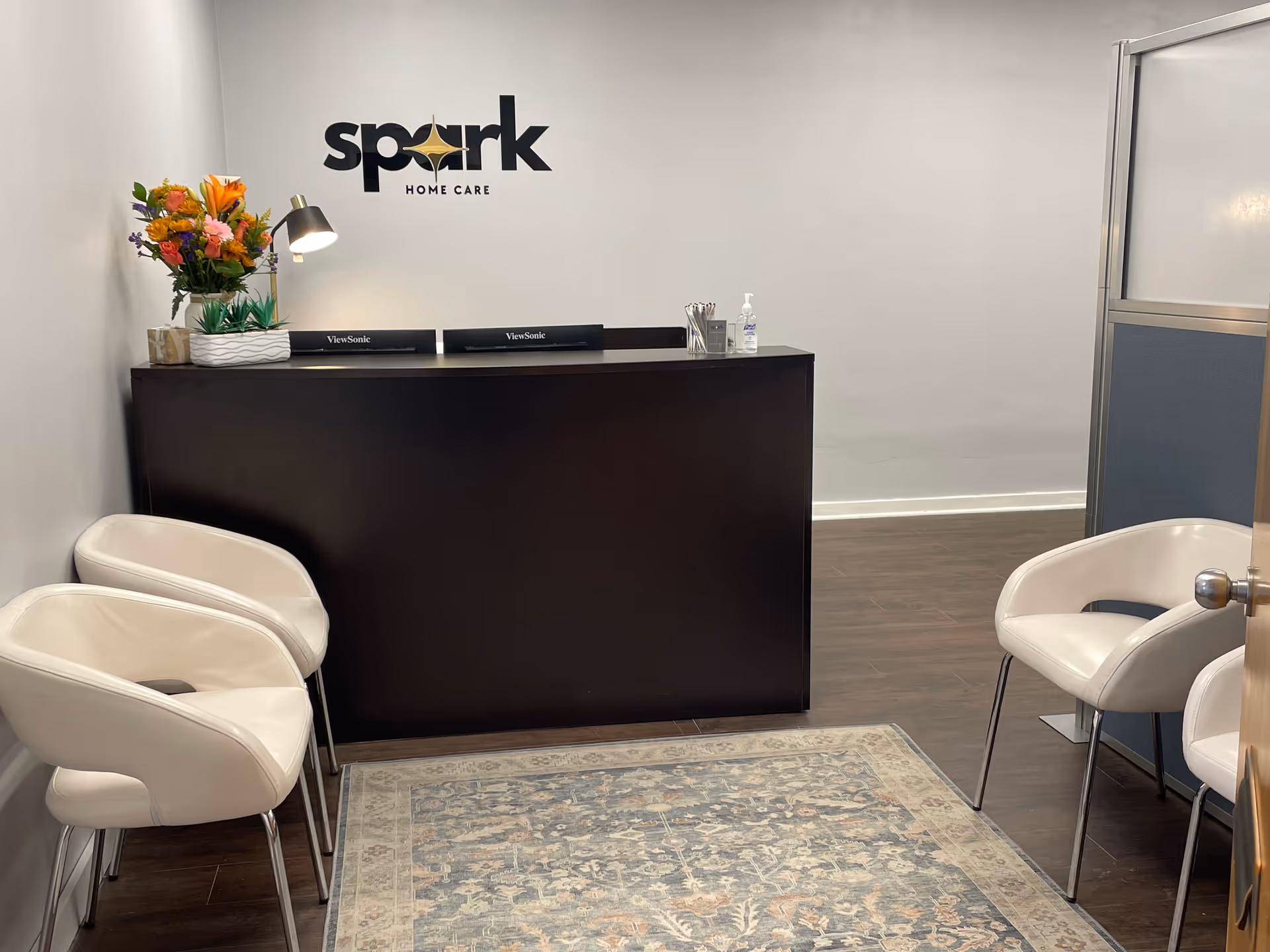 Reception area featuring a dark front desk, flowers, white chairs, and Spark Home Care sign.