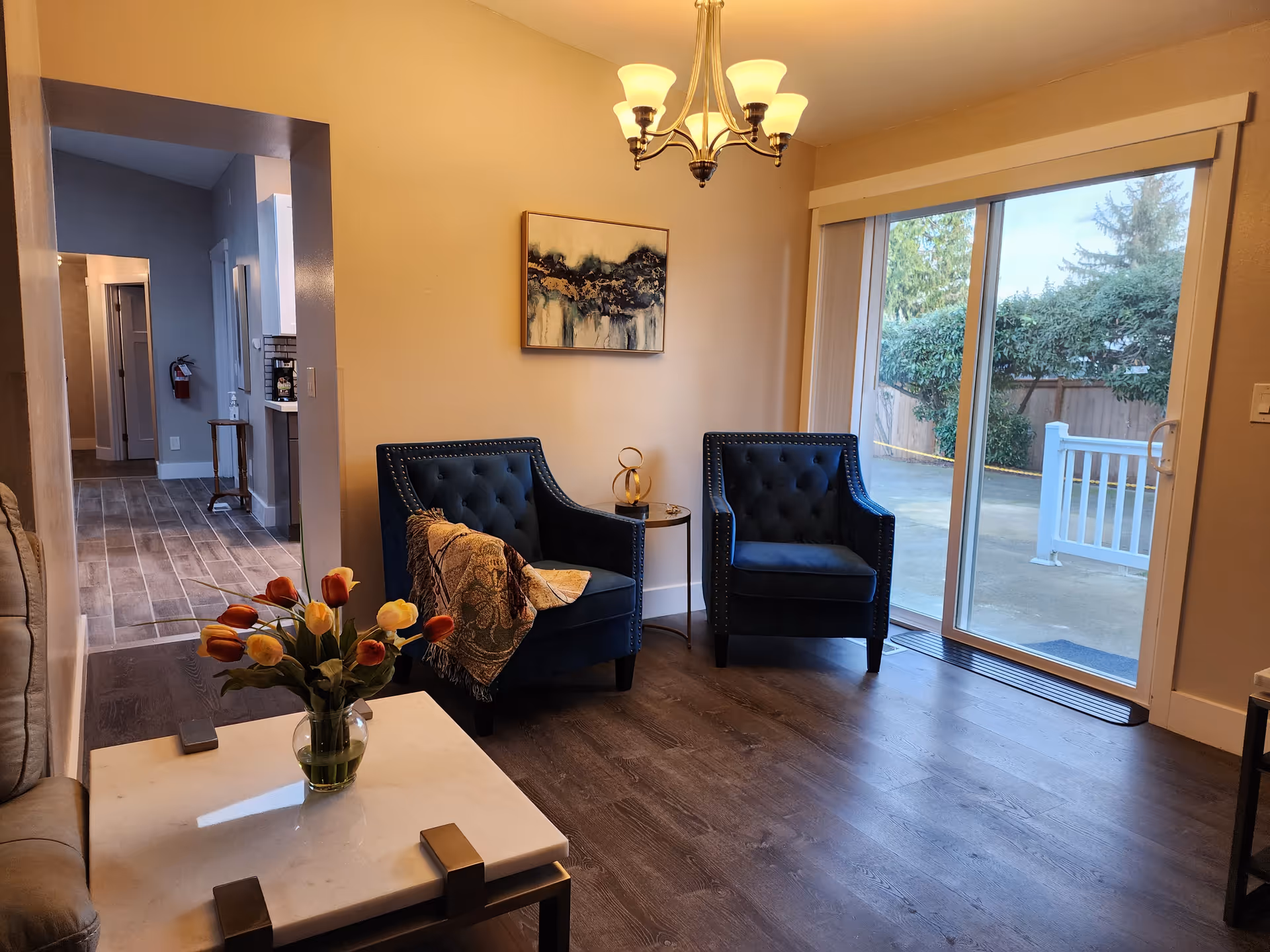 Cozy living room with two blue chairs, flowers, coffee table, and sliding door to outdoors.