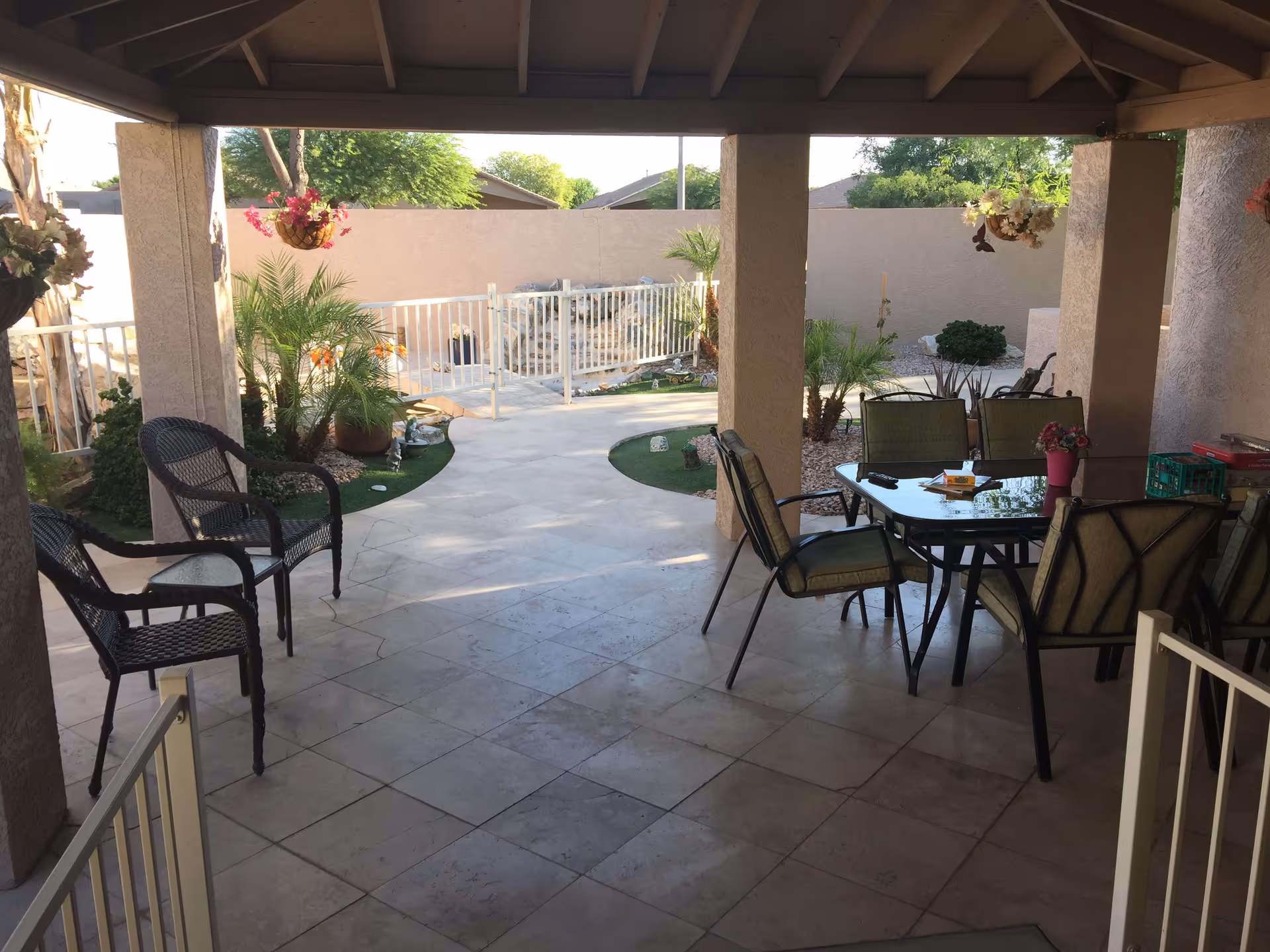 Covered outdoor patio area with tiled floor, featuring a glass-top table surrounded by six cushioned chairs on the right side and two black wicker chairs on the left side. The patio overlooks a fenced garden area with various plants, small palm trees, and hanging flower baskets. The background shows a beige wall and some trees beyond it.