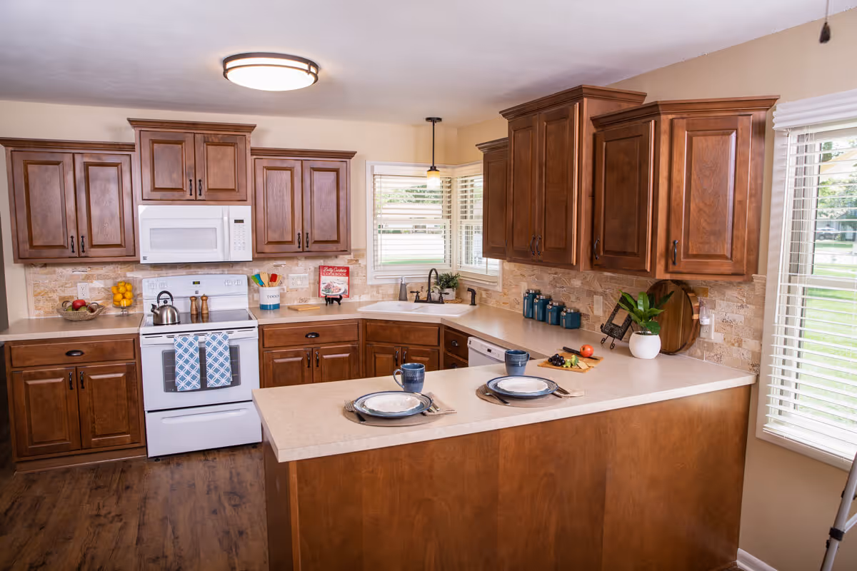 Kitchen with warm wooden cabinets, an island set for two, a white stove and microwave, and windows with blinds.