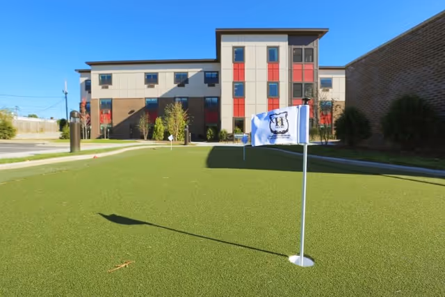 A putting green with a flag in front of a modern three-story senior living building under a clear blue sky.
