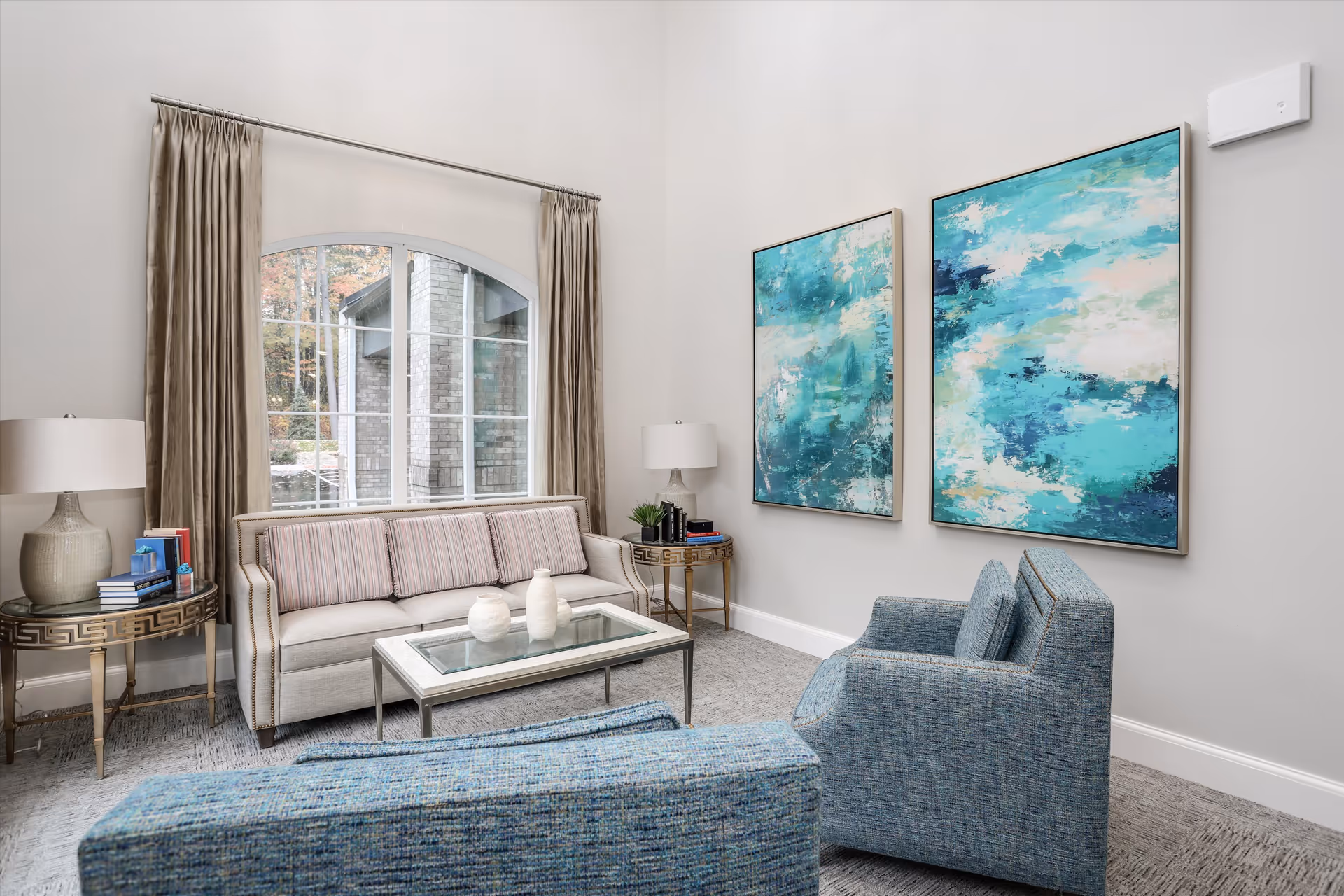 A modern living room with a beige sofa, two blue armchairs, a glass coffee table with decorative vases, two side tables with lamps and books, large window with beige curtains, and two abstract blue and white paintings on the wall.