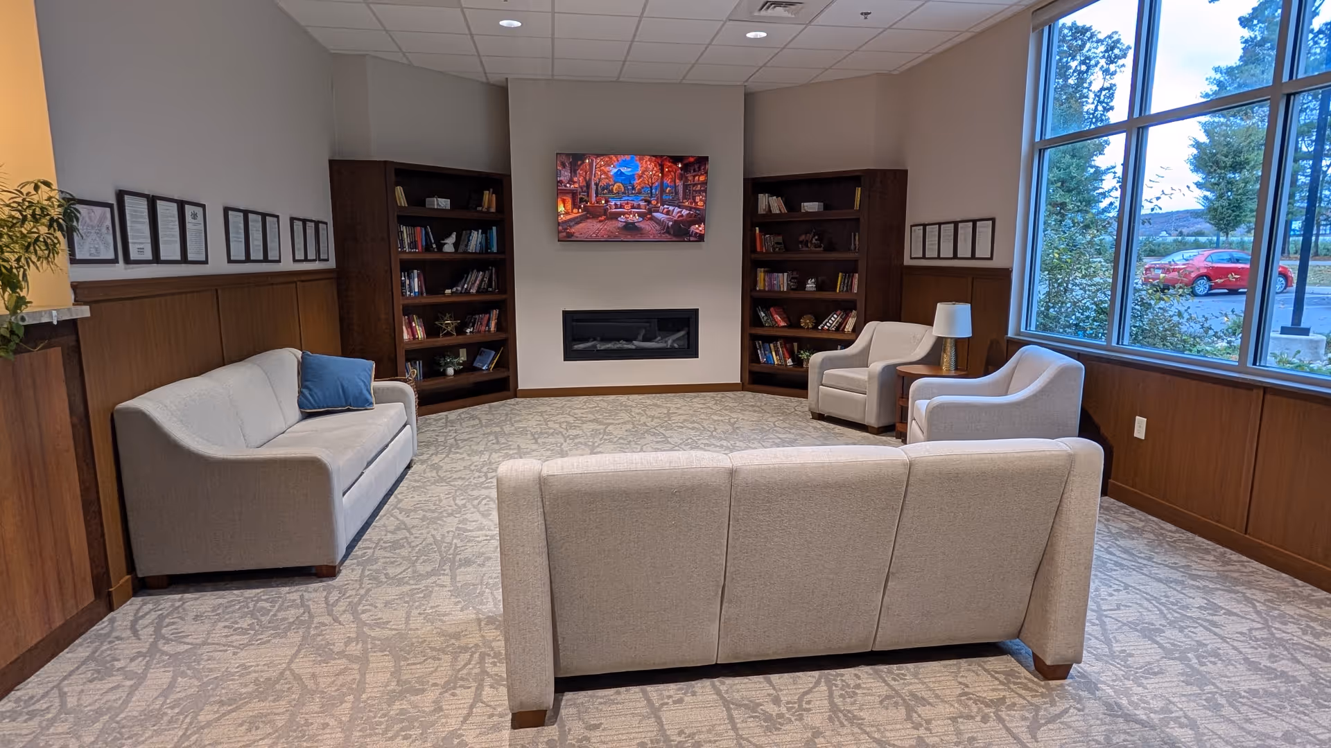 Cozy senior living lounge with beige sofa set, bookshelves, fireplace, and large windows.