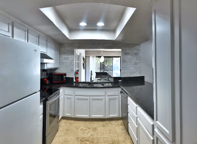 Modern kitchen with white cabinetry, black countertops, stainless steel appliances, a sink facing a pass-through window, and a recessed skylight.