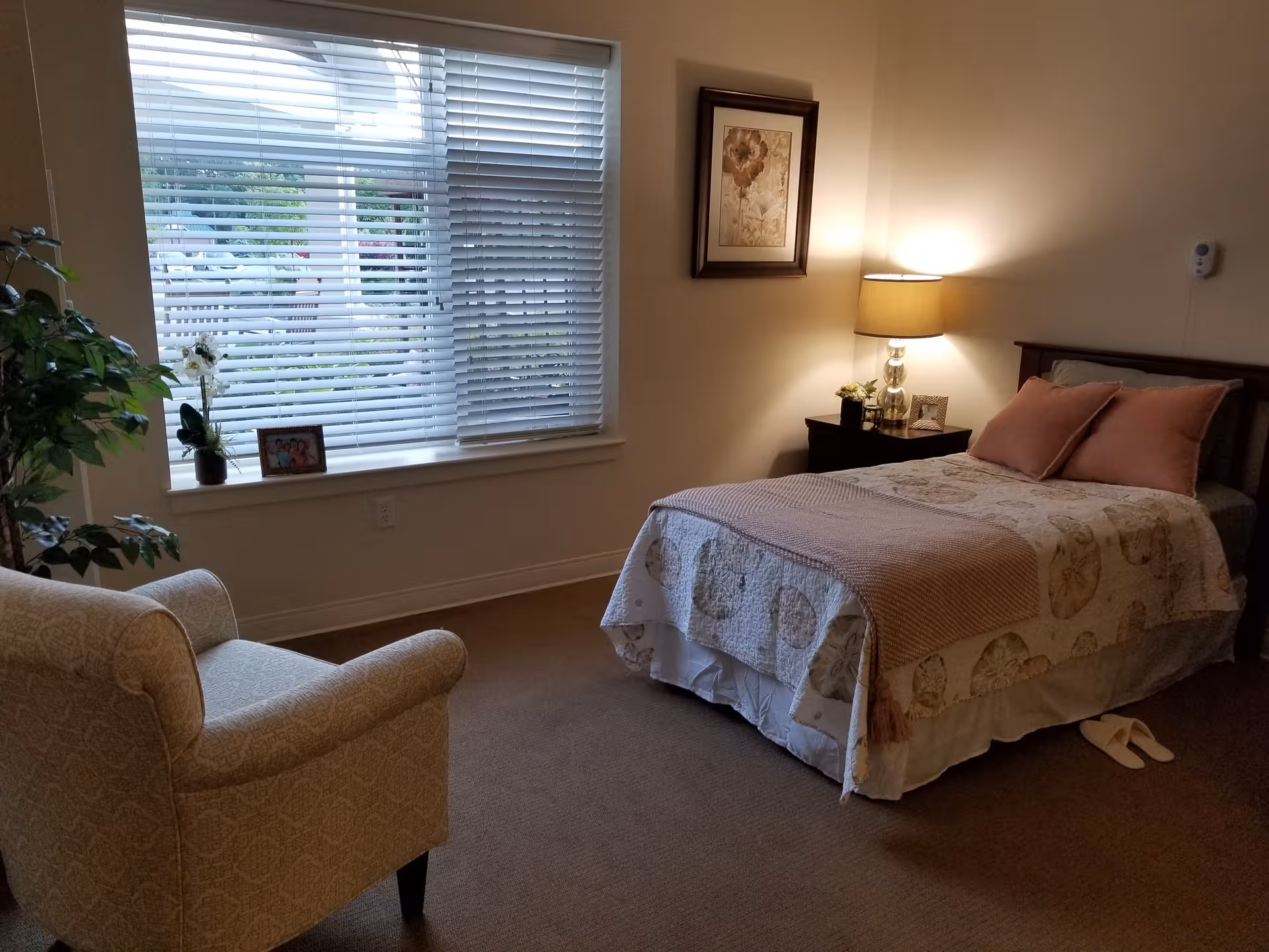 Senior living bedroom with bed, nightstand, lamp, chair, window blinds, and slippers.