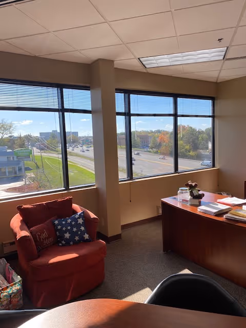 Office area with red chair, wooden desk, papers, and large windows overlooking outdoors.