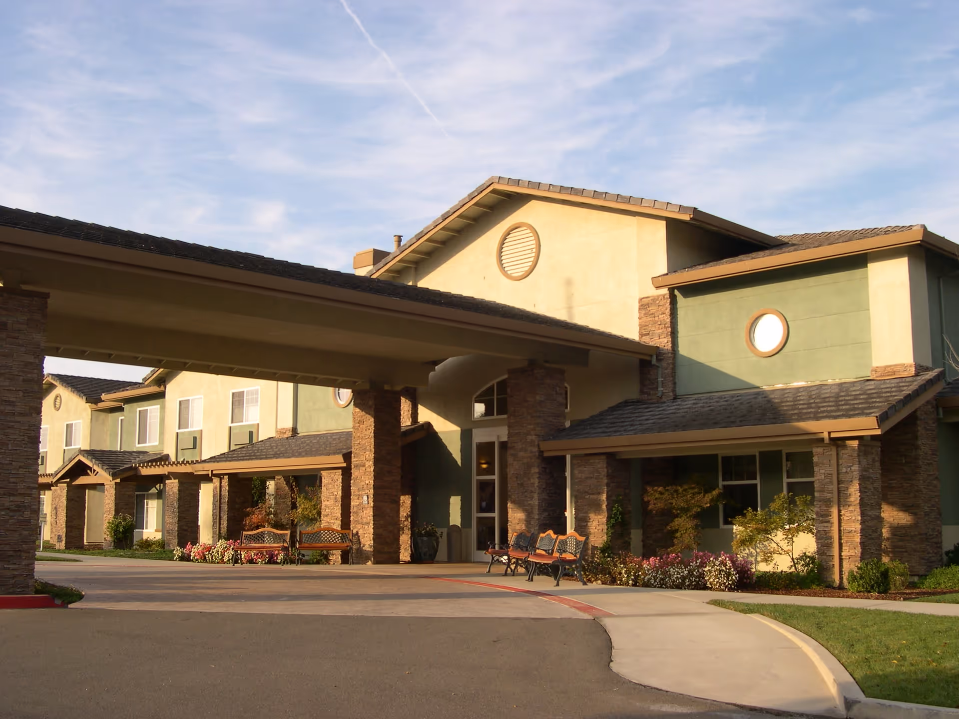 Exterior view of Eskaton Granite Bay senior living facility showing the main entrance with a covered driveway, stone pillars, benches, and landscaped flower beds under a partly cloudy sky.