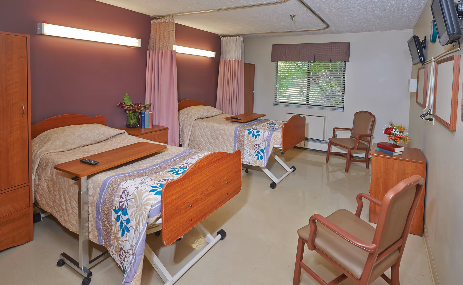 A senior living facility bedroom with two hospital-style beds, each with a wooden headboard and footboard, beige bedspreads, and floral blankets. There are two chairs, a small wooden cabinet with books and flowers, a window with blinds and a valance, and two wall-mounted televisions. The walls are painted in a combination of purple and beige, and there are overhead lights above each bed.