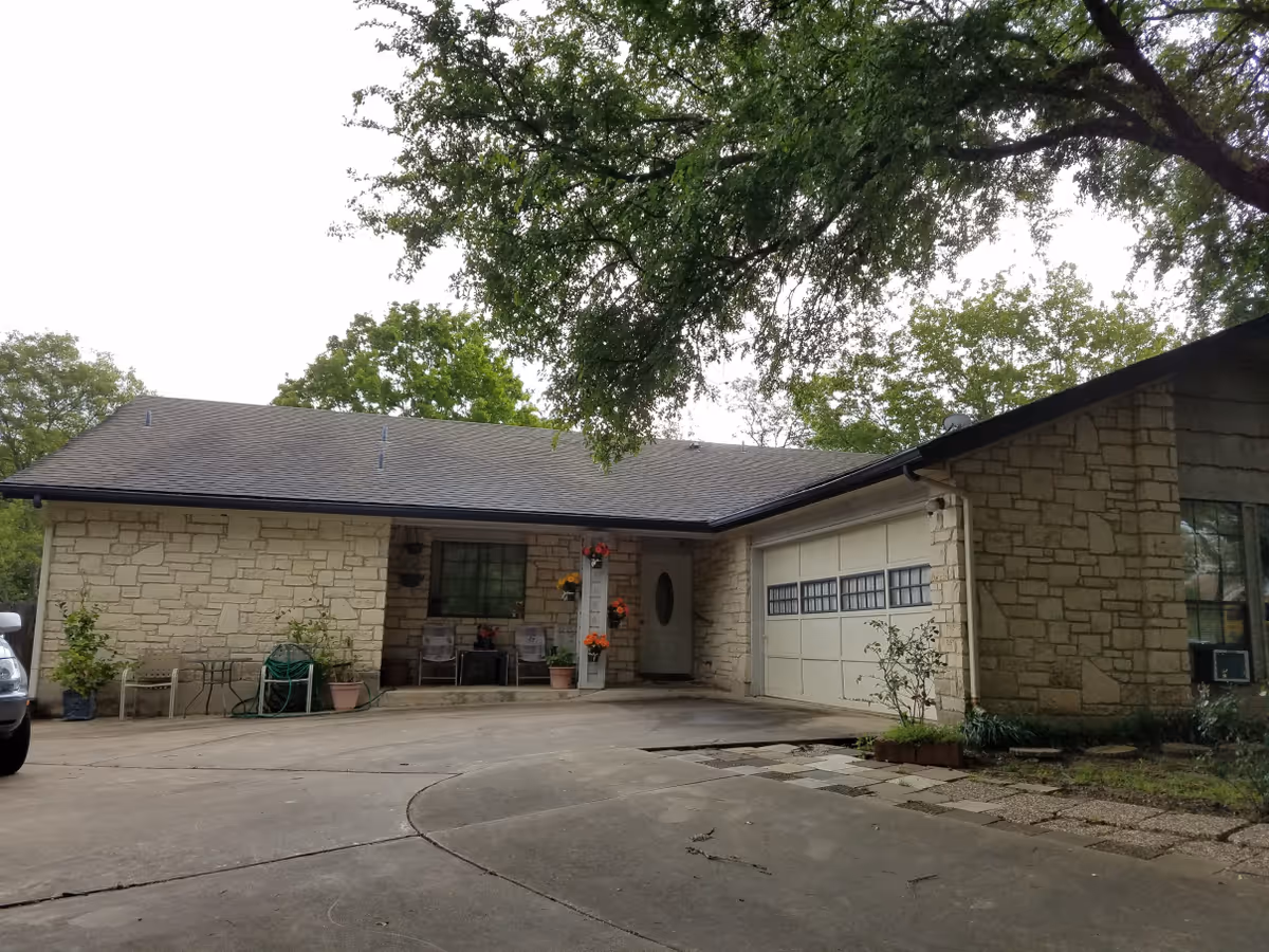 Exterior view of a single-story stone building with a garage door, a small porch with chairs and potted plants, and a large tree overhead. The driveway is concrete and there are some plants and garden hose near the porch.