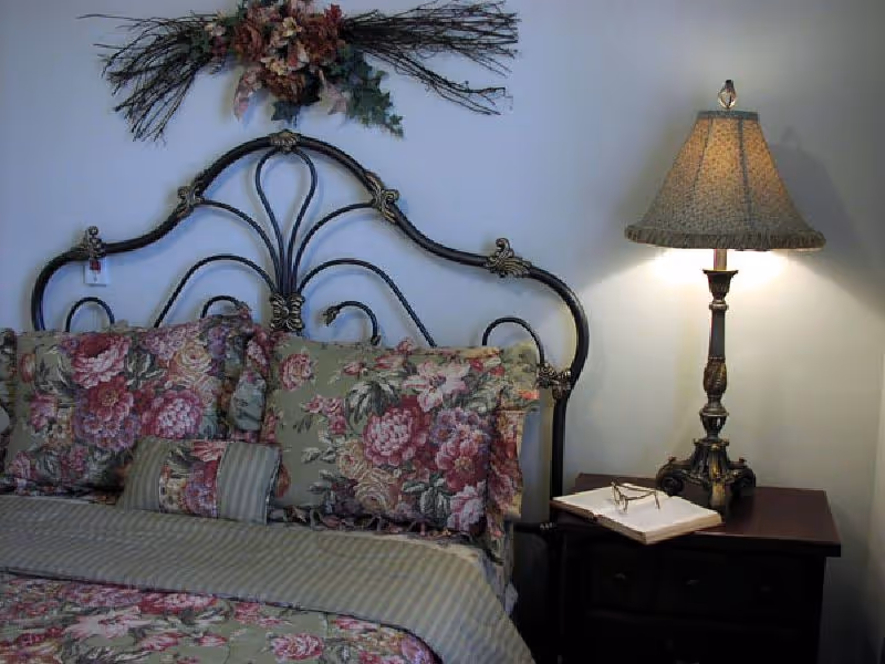Decorative metal headboard with floral bedding and pillows beside a nightstand holding a lamp and book.