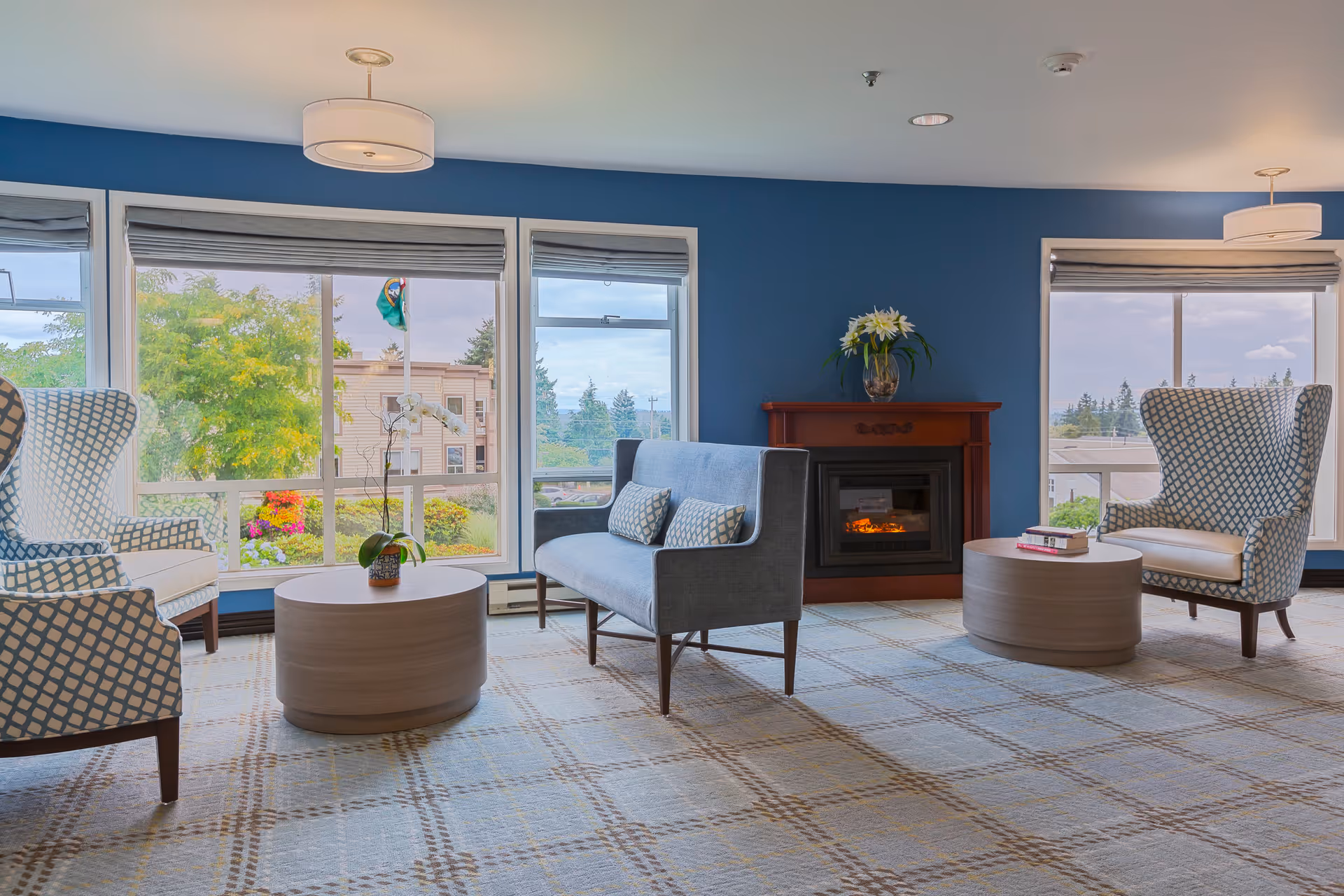 A cozy living room area with two patterned armchairs, a blue loveseat with matching pillows, two round wooden coffee tables, and a fireplace with a flower vase on top. Large windows with gray blinds provide a view of trees and buildings outside. The walls are painted blue and the carpet has a plaid pattern.