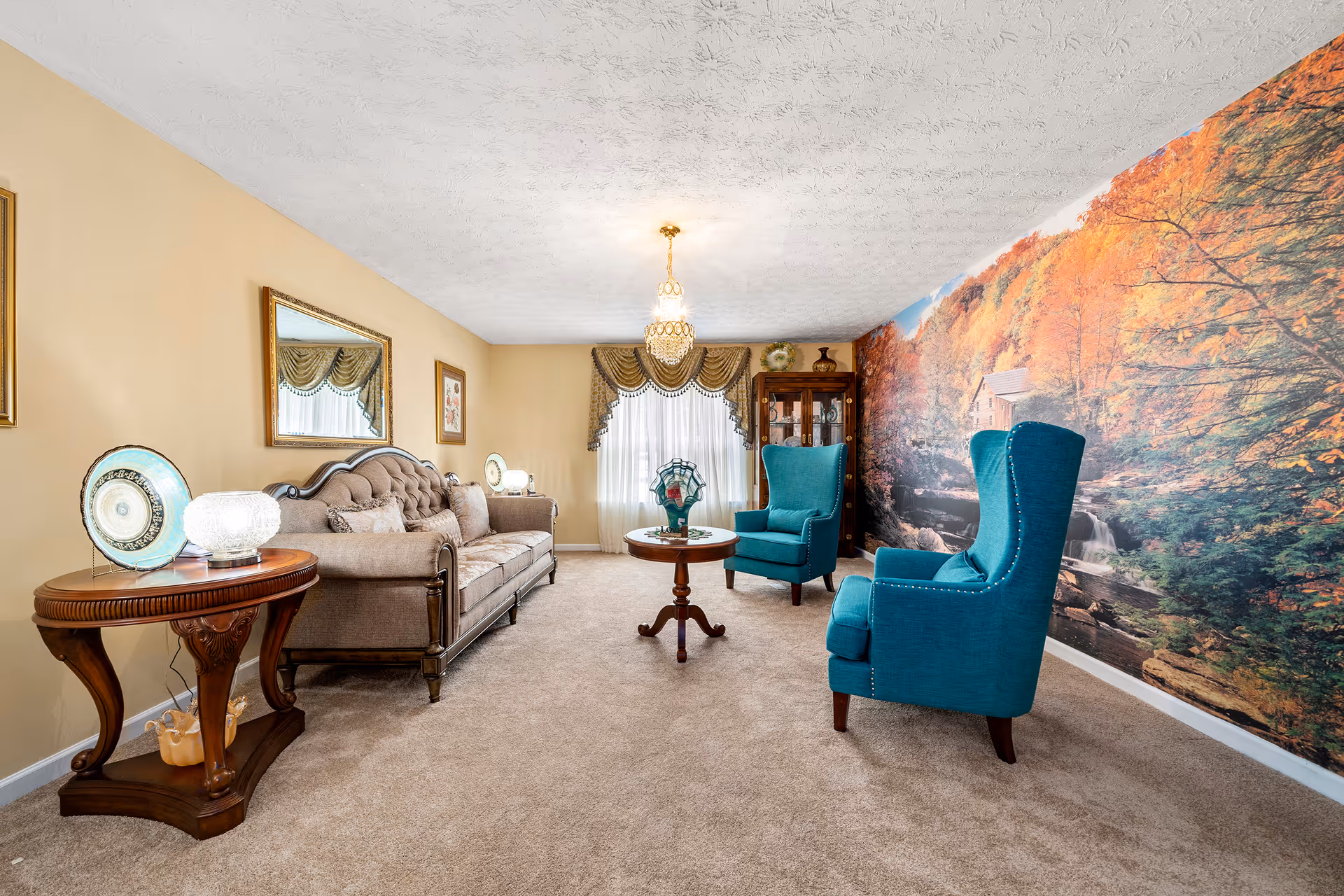 A cozy living room with beige sofa, turquoise chairs, mural wall, and chandelier.