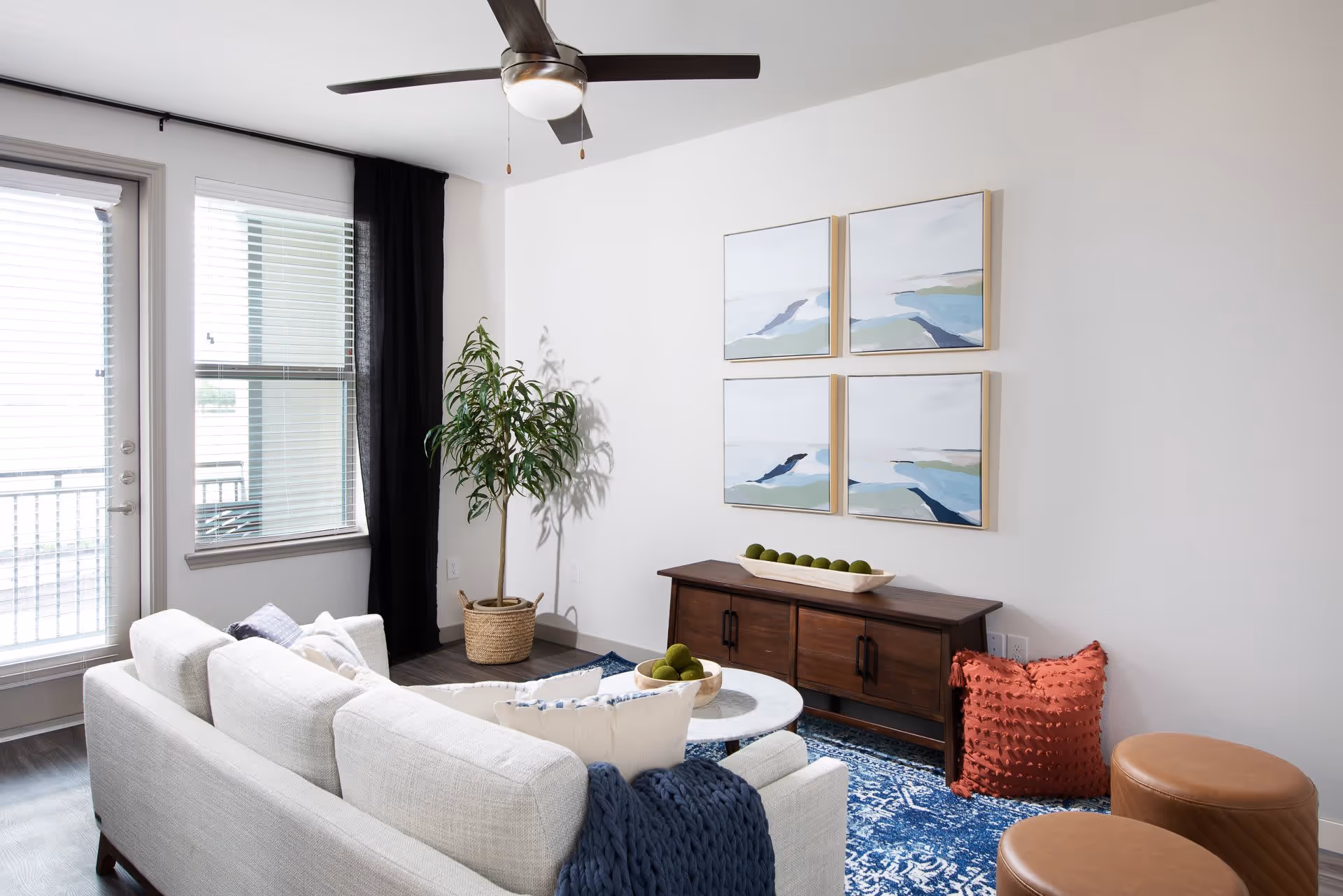 A modern living room with a light gray sofa adorned with pillows and a blue knitted throw. In front of the sofa is a round white marble coffee table with decorative green balls. Behind the coffee table is a dark wooden console table with a long white bowl filled with green decorative balls. Four abstract landscape paintings are hung on the white wall above the console. To the left, there is a large window and a glass door with blinds and a black curtain. A potted plant in a woven basket sits in the corner. Two brown leather ottomans are placed on a blue patterned rug. A ceiling fan with a light fixture is mounted on the white ceiling.
