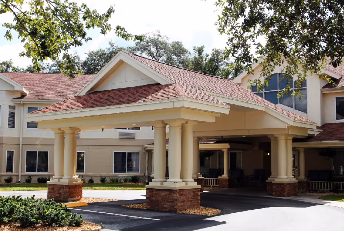 Entrance of The Springs at Lady Lake Assisted Living facility featuring a covered driveway with four large columns supporting a peaked roof, surrounded by a beige building with multiple windows and greenery.