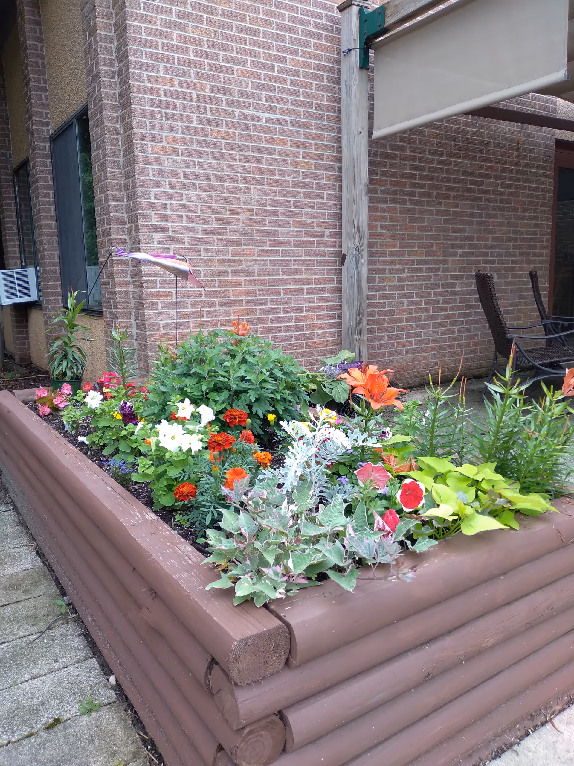 Exterior brick wall with a raised planter bed of colorful flowers and patio seating.