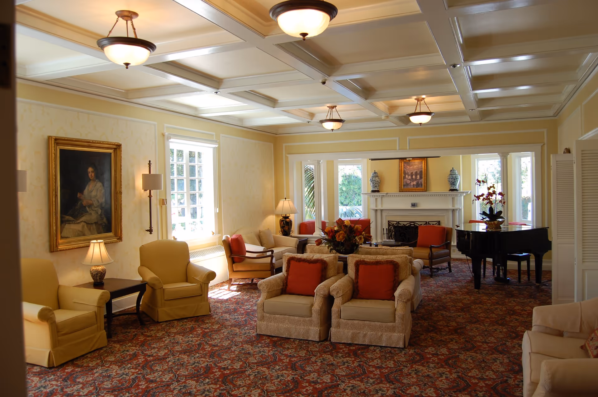 Bright, elegant common room with upholstered armchairs and sofas, a fireplace, grand piano, and coffered ceiling.
