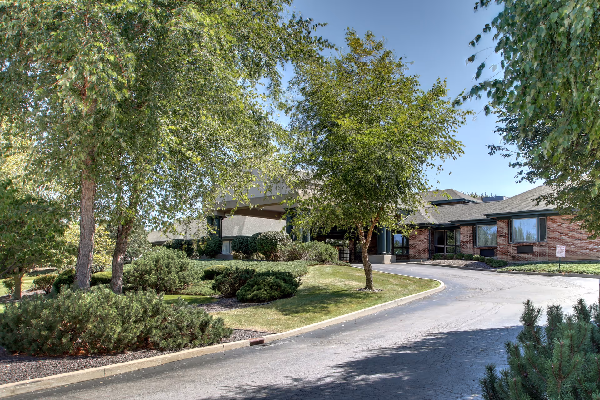 Exterior view of Regency Care facility showing a curved driveway leading to the entrance, surrounded by green trees, shrubs, and well-maintained landscaping under a clear blue sky.