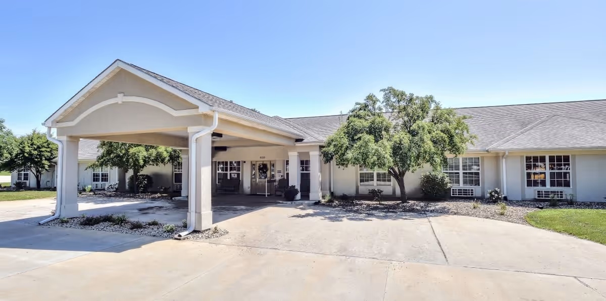 Front exterior view of a single-story senior living facility building with a covered entrance, several windows, and a tree in front. The sky is clear and blue.