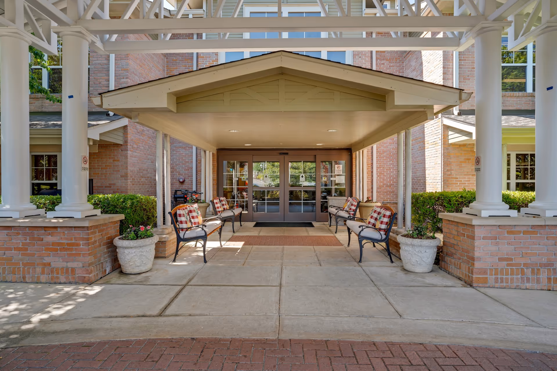 Covered main entrance of a brick building with white columns, cushioned benches, potted plants, and glass doors.