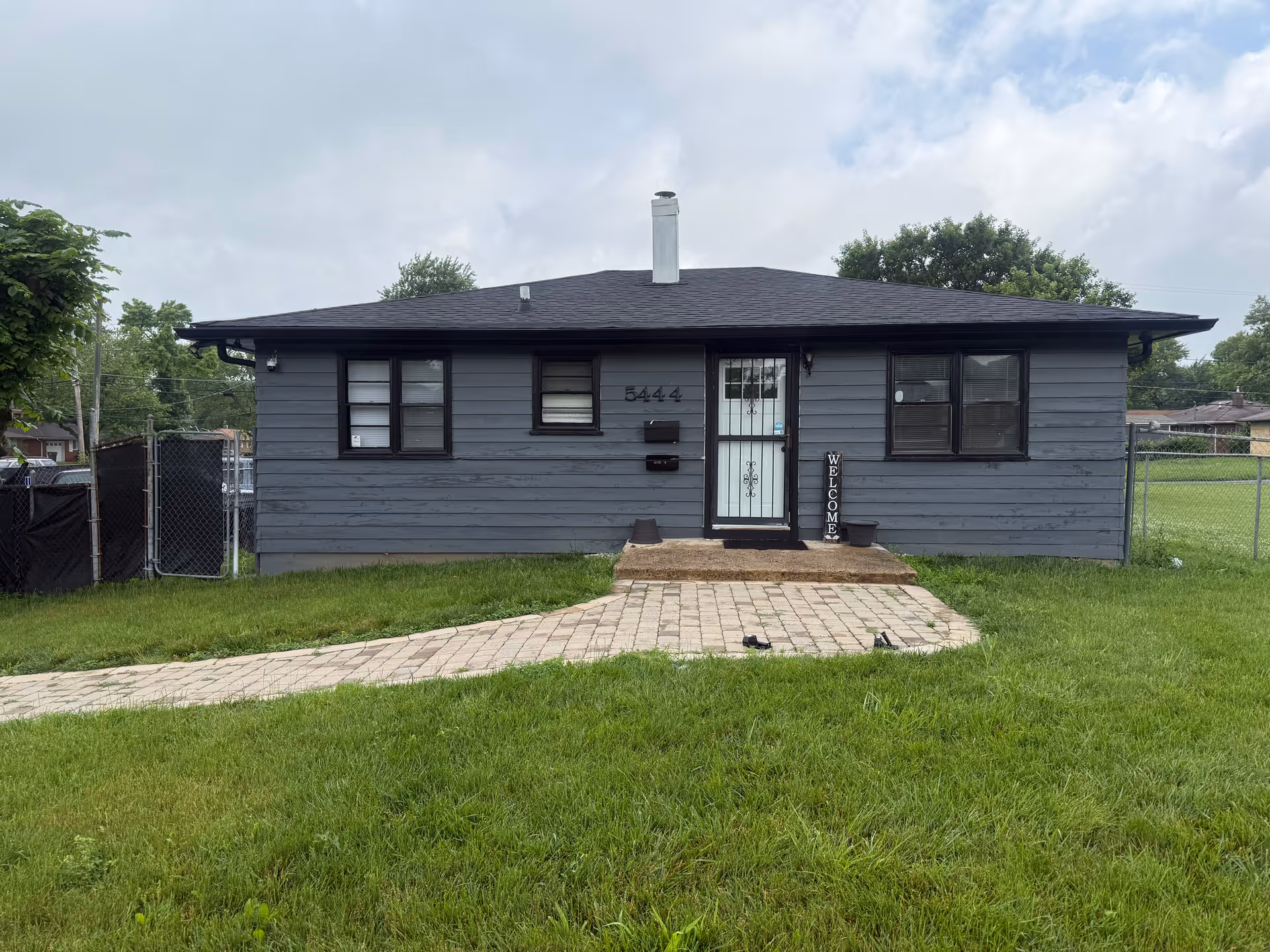 Small blue-gray single-story house with a paved walkway and welcome sign in front yard.
