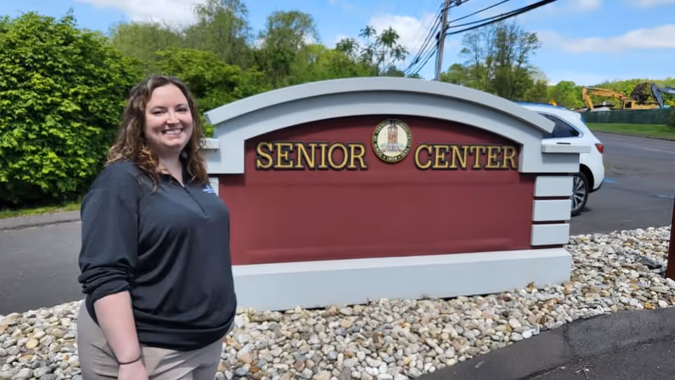 Smiling woman stands beside a large red Senior Center sign outdoors.