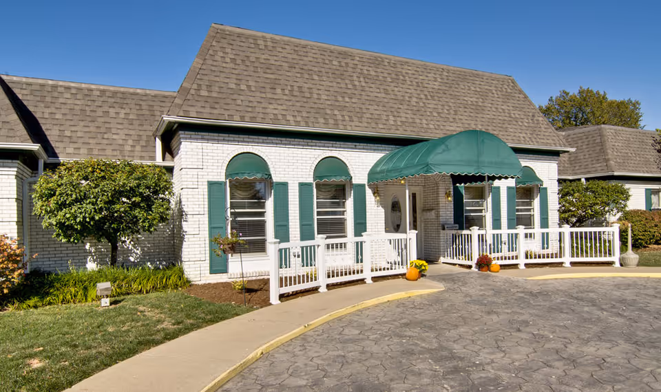 Exterior view of a single-story building with white brick walls, green window shutters, and a green awning over the entrance. There is a paved driveway and a sidewalk leading to the entrance, with some landscaping including bushes and small trees around the building.