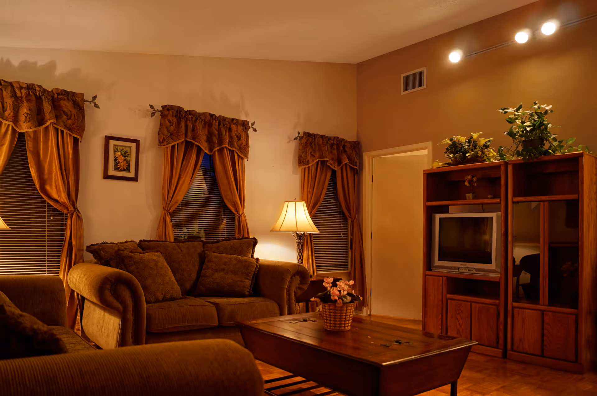 A cozy living room with brown upholstered sofas, a wooden coffee table with a flower basket, a wooden entertainment center with a TV, and three windows with brown curtains. A floor lamp provides warm lighting, and there are plants on top of the entertainment center.