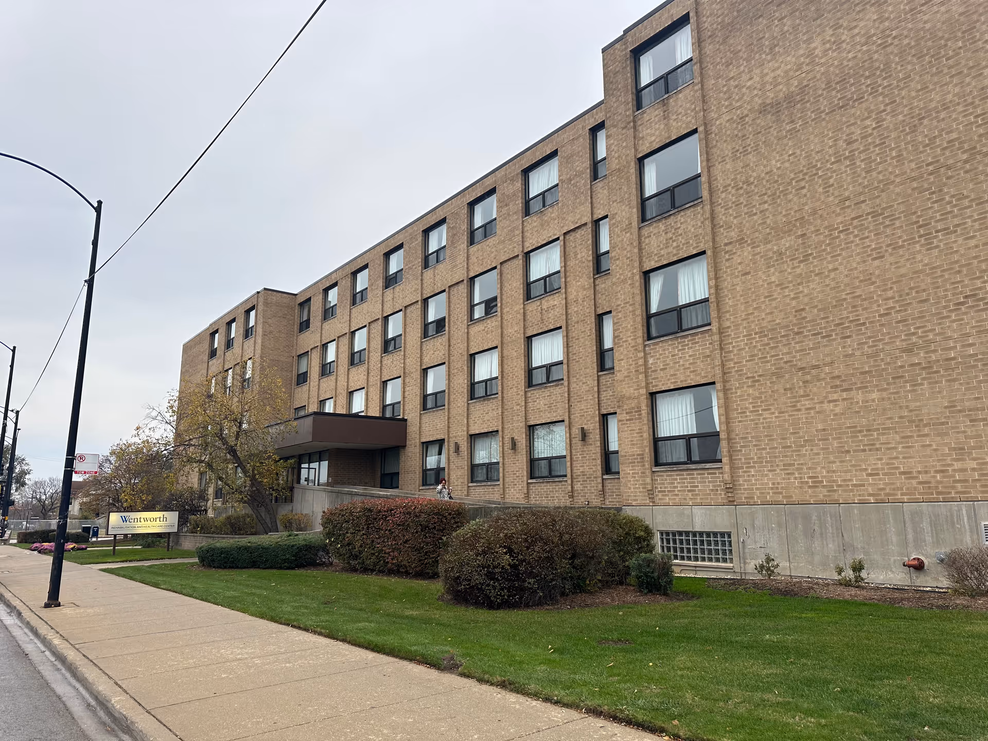 Four-story brick senior care building with an entrance canopy, lawn and a 'Wentworth' sign near the sidewalk.