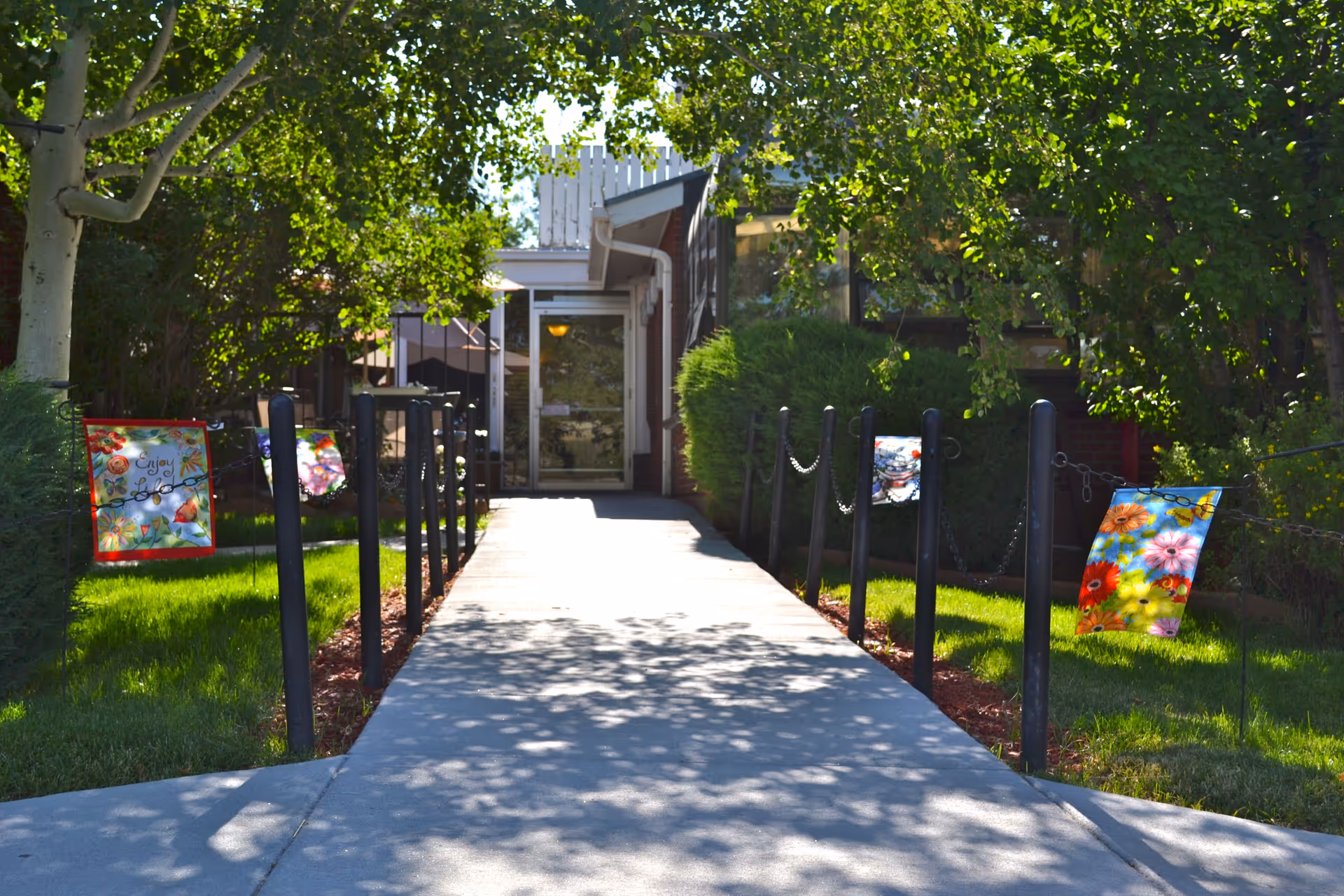 Tree-shaded walkway leads to the Wheatridge Manor Care Center entrance, with colorful posters along posts.
