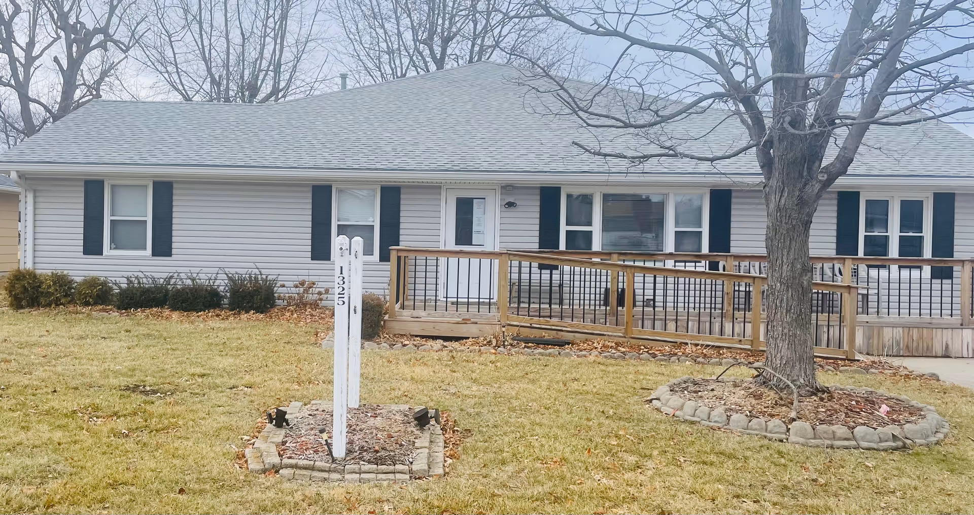 A single-story residential care building with a wooden ramp, tree, and front yard.