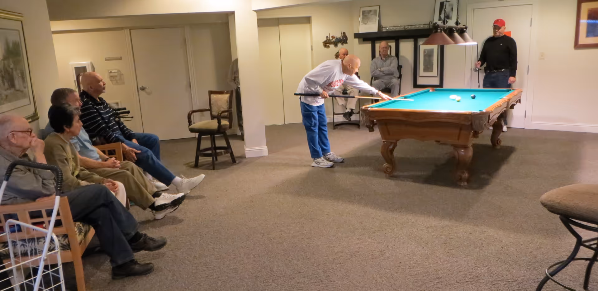 A group of elderly people in a common room at Plaza At Highlands Crossing. Two men are playing pool on a billiards table while several others sit on chairs watching the game. The room has beige walls, carpeted floor, and framed artwork on the walls.