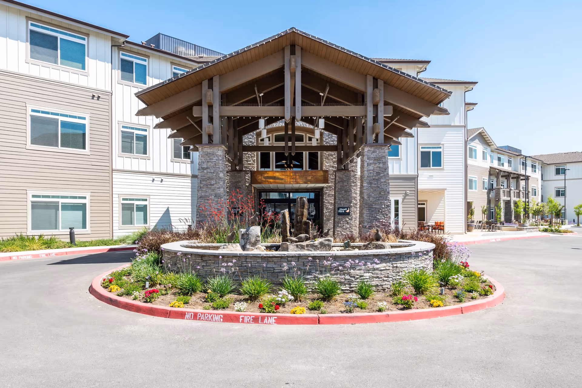 Front exterior view of Quail Park at Shannon Ranch facility showing a covered entrance with stone pillars, a circular landscaped area with flowers and rocks, and a clear blue sky.