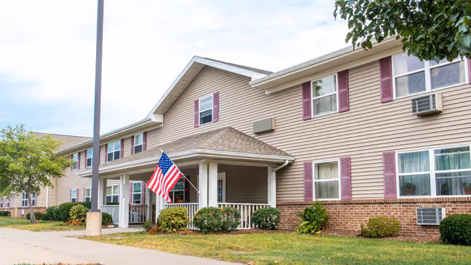 Exterior view of a beige senior living facility with purple shutters and a flag