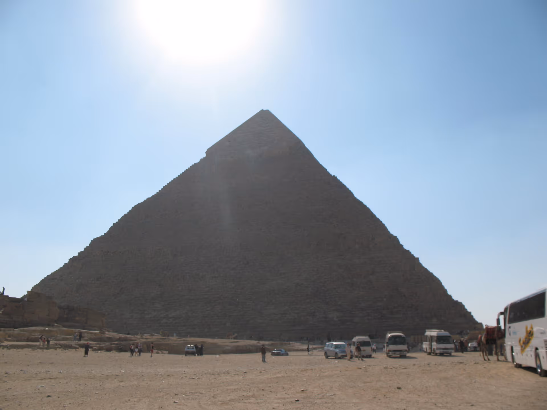 Giant pyramid under bright sun with tourists and parked buses in a desert.