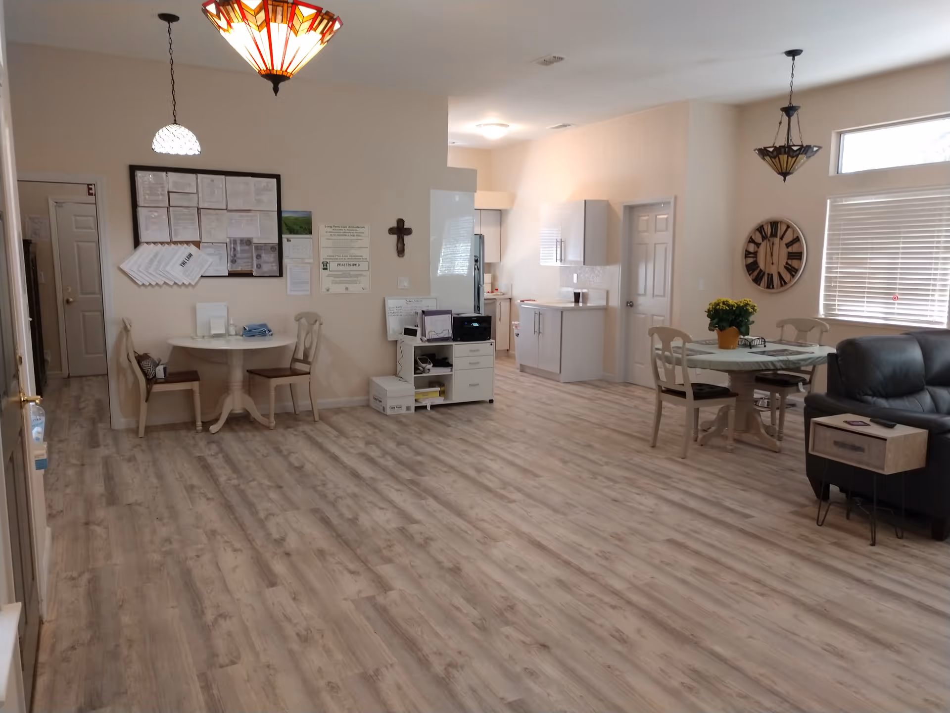 Interior view of a senior living facility common area with light wood flooring, two small round dining tables with chairs, a black leather sofa, a large wall clock, and a small kitchen area in the background. The walls are light-colored, and there are three hanging light fixtures with decorative shades. Various notices and a cross are mounted on the wall near one of the tables.
