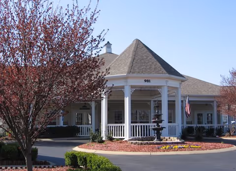 Front exterior of a senior living facility with white porch, gazebo, fountain, and landscaped circular drive.