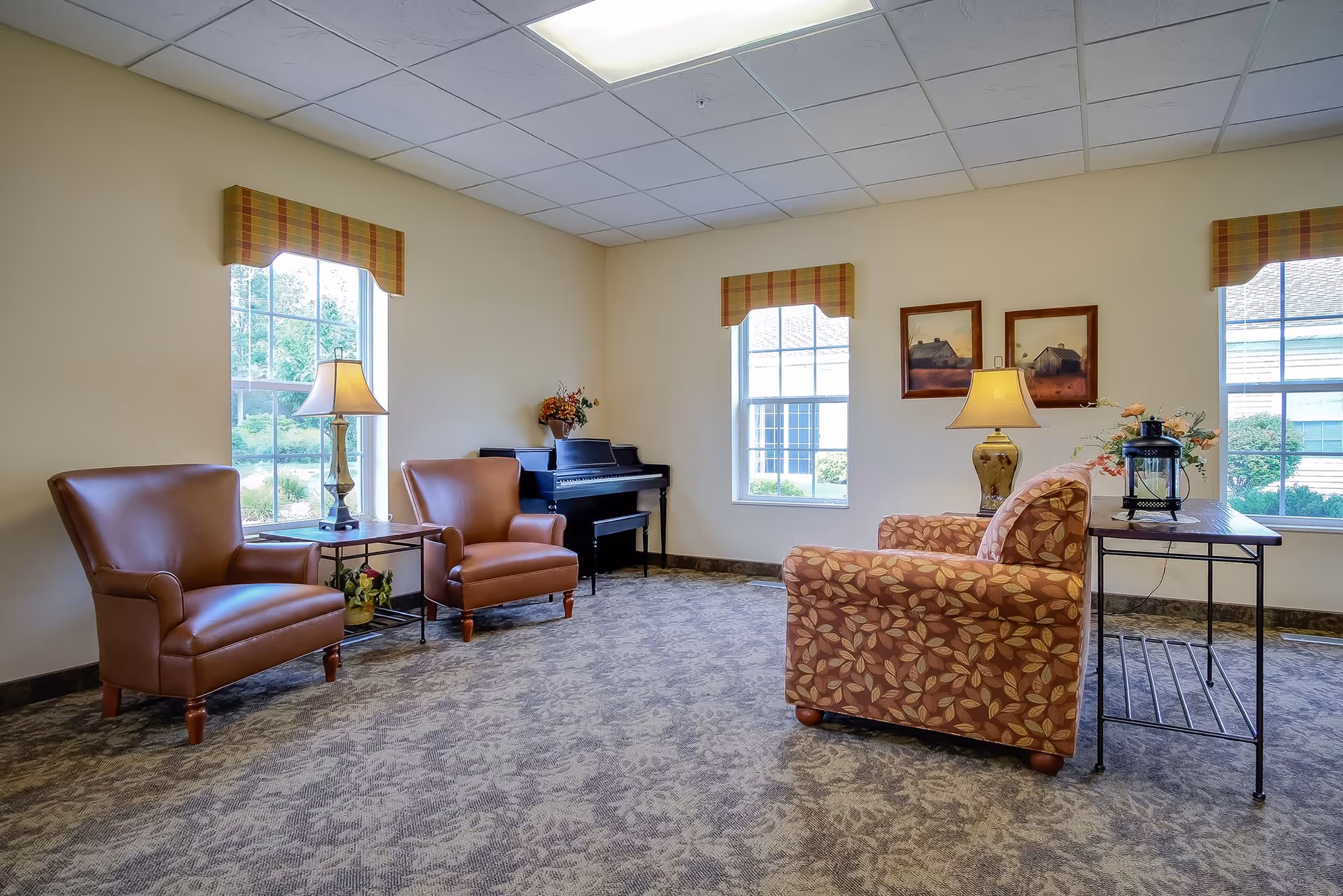 Assisted living lounge with two brown armchairs, a floral-pattern chair, piano, lamps, and windows.