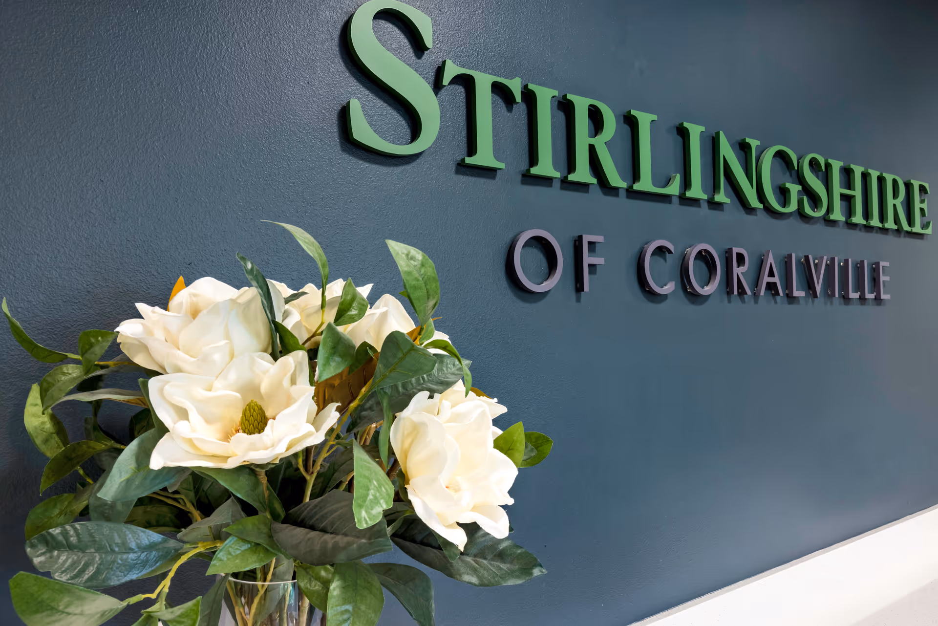 Close-up view of a dark blue wall with the words 'Stirlingshire of Coralville' mounted on it in green and gray letters, accompanied by a vase of white flowers with green leaves in the foreground.